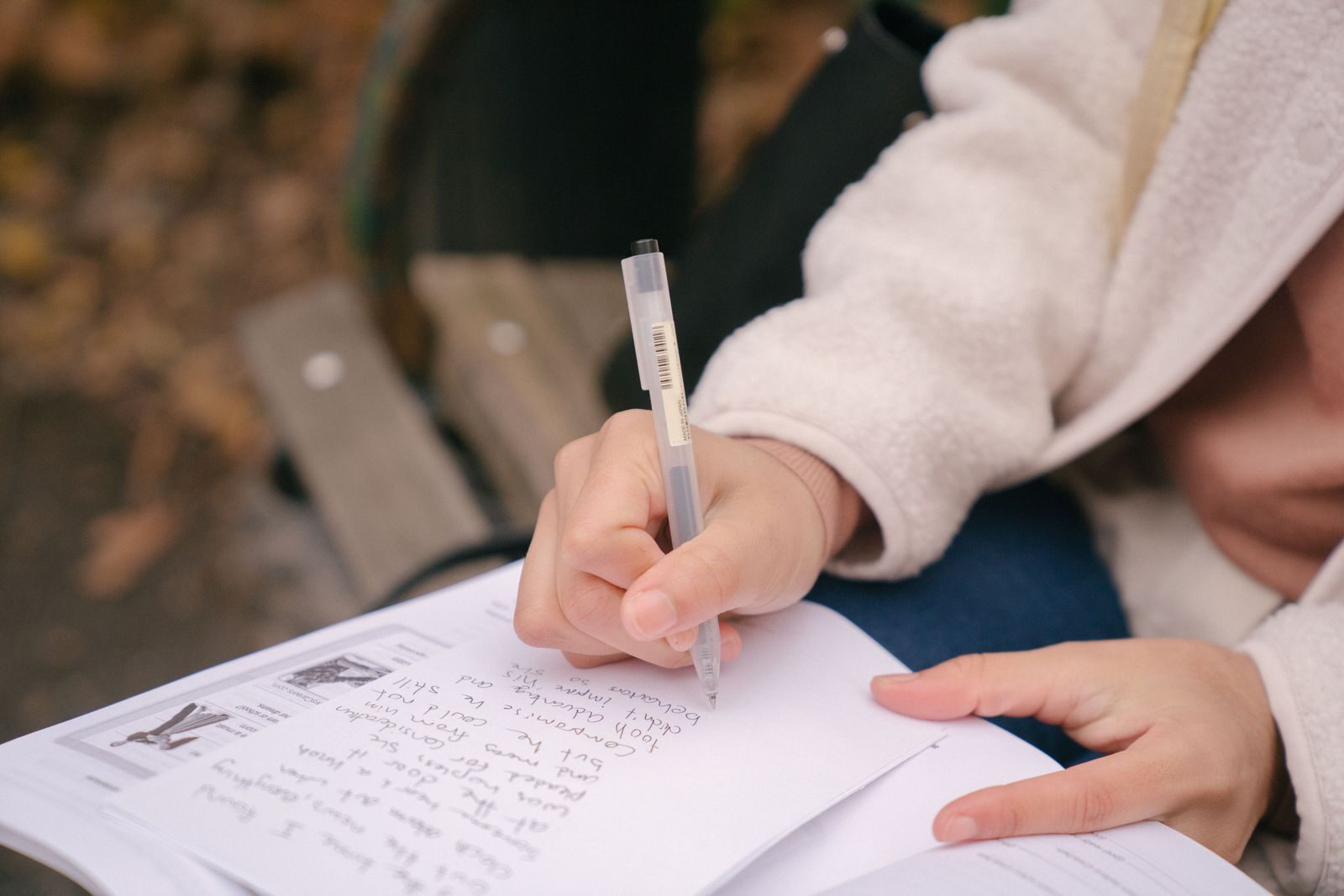 Crop unrecognizable woman in casual outfit sitting on wooden bench while taking notes on paper near textbook in street with foliage on ground in daylight in autumn